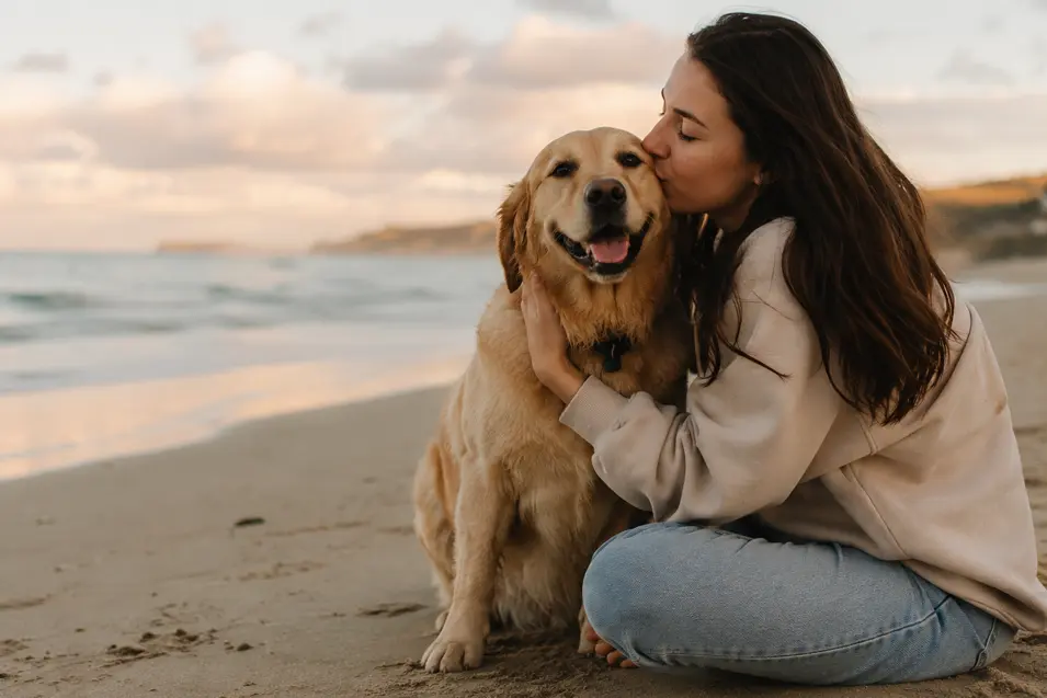 A woman cuddles her happy dog on a peaceful beach at sunset, sharing a tender moment beside the ocean.