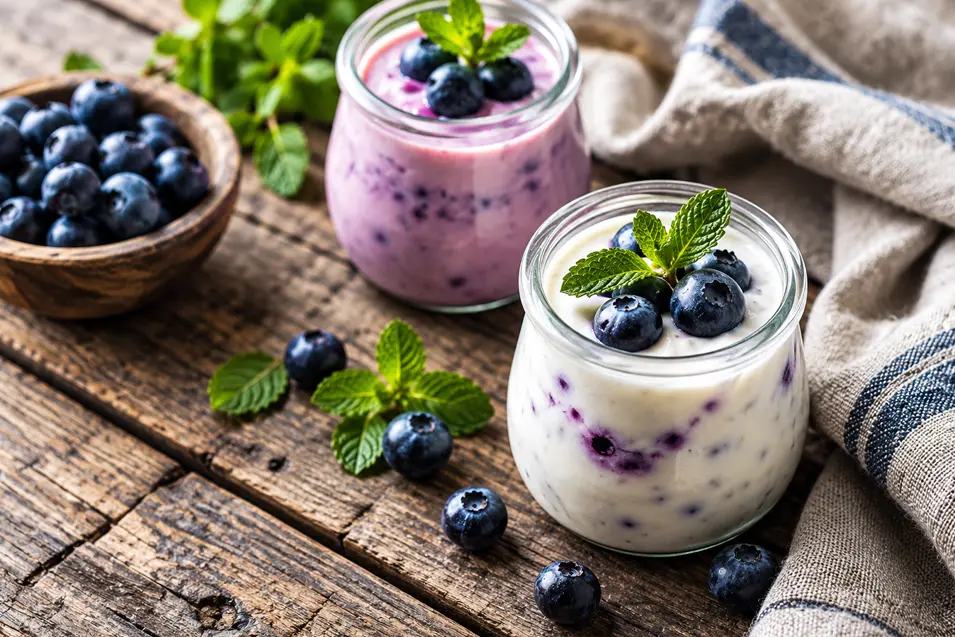 Two jars of creamy blueberry yogurt topped with fresh blueberries and mint sit on a rustic wooden table beside a bowl of berries.