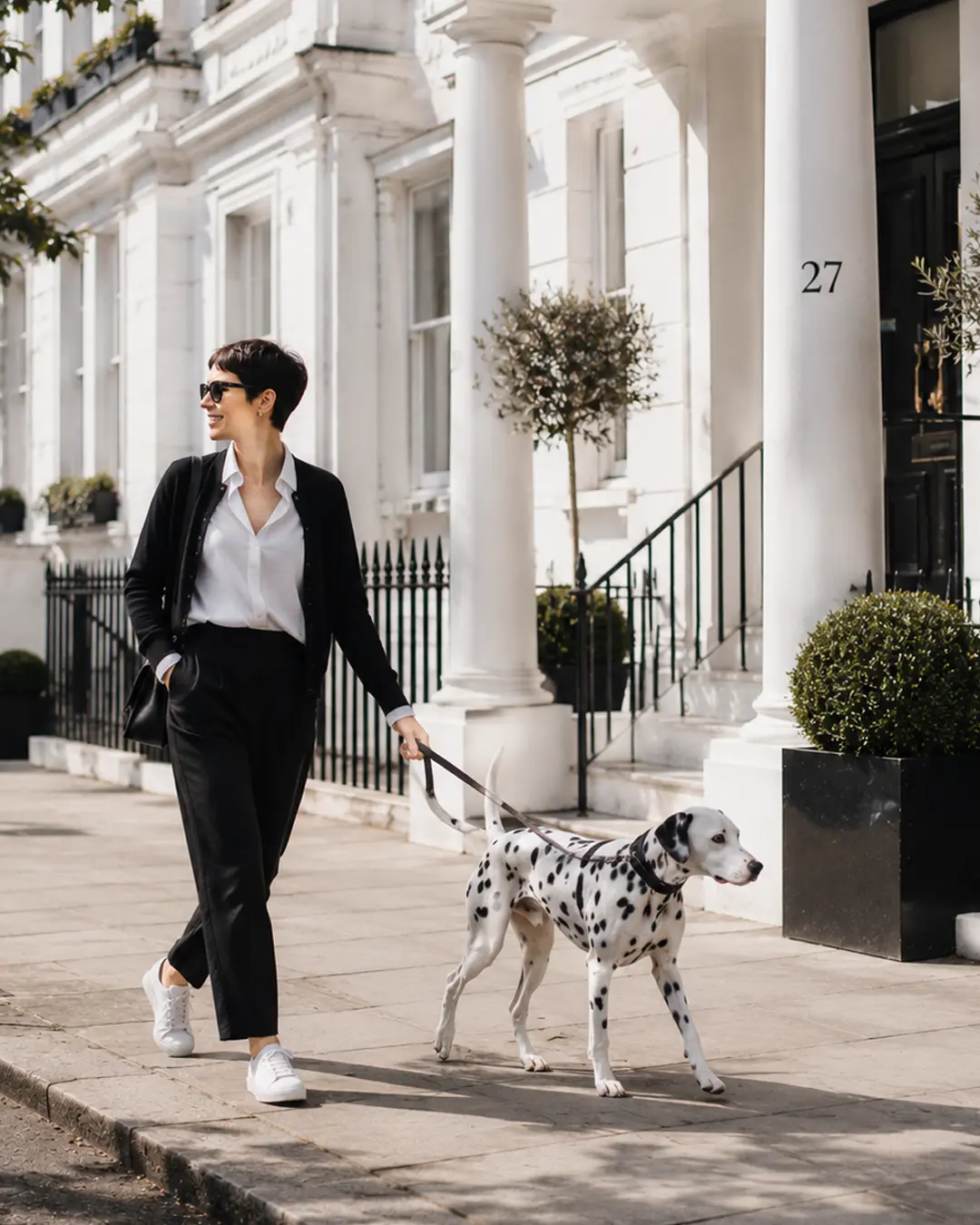 A stylish woman in classic black and white walks her Dalmatian along an elegant, sunlit city street lined with white townhouses.