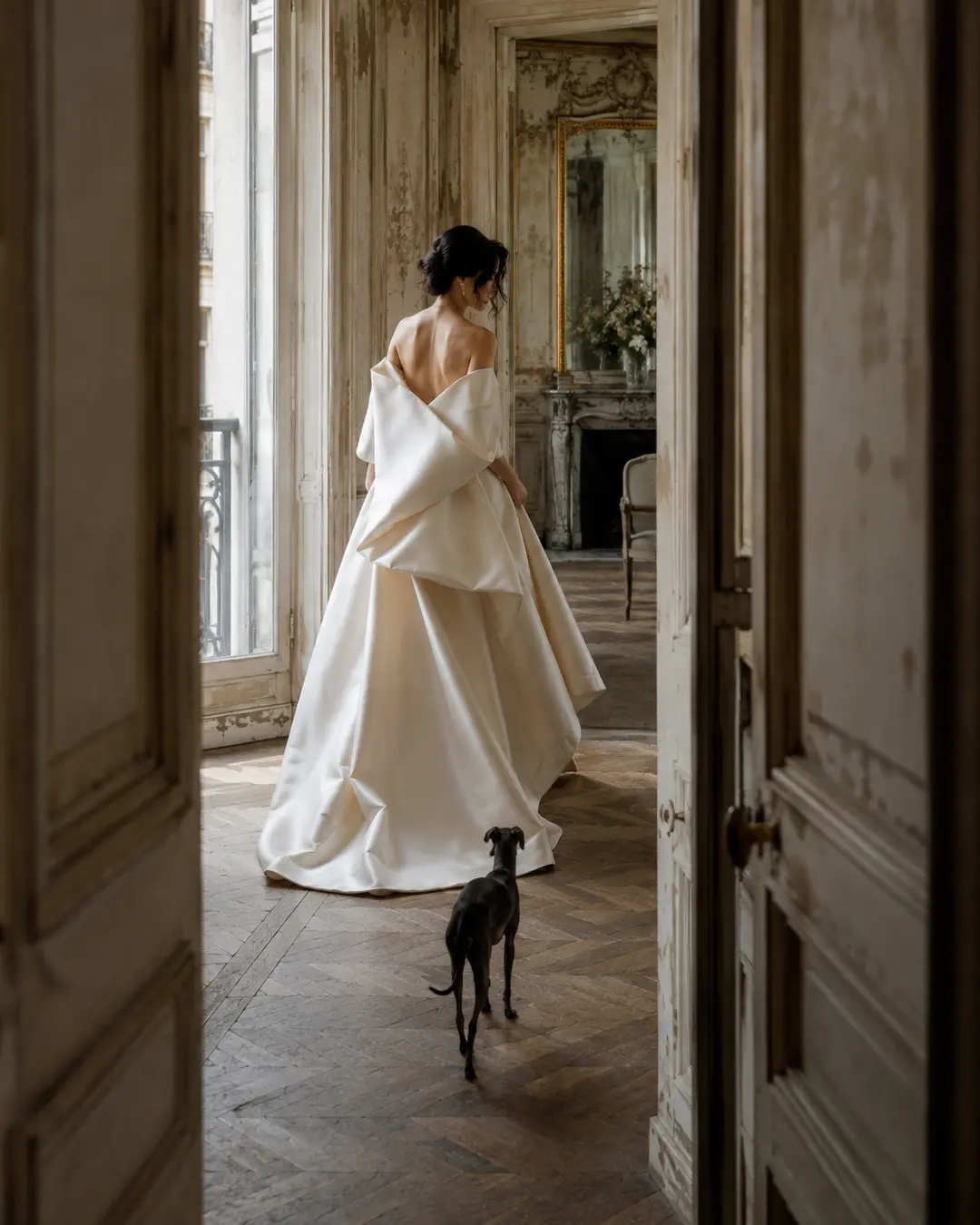 A bride in a flowing white gown walks through a faded Parisian interior, followed by a small black dog in a quiet, elegant wedding moment.