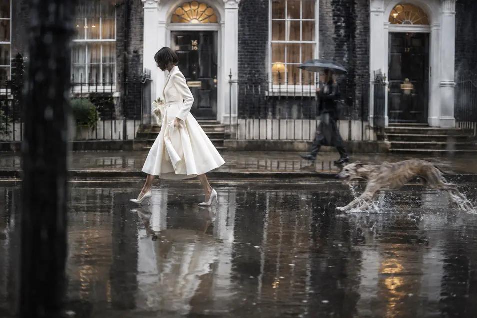 A bride in a chic white coat dress crosses a rain-soaked London street while her playful dog races through puddles beside her.