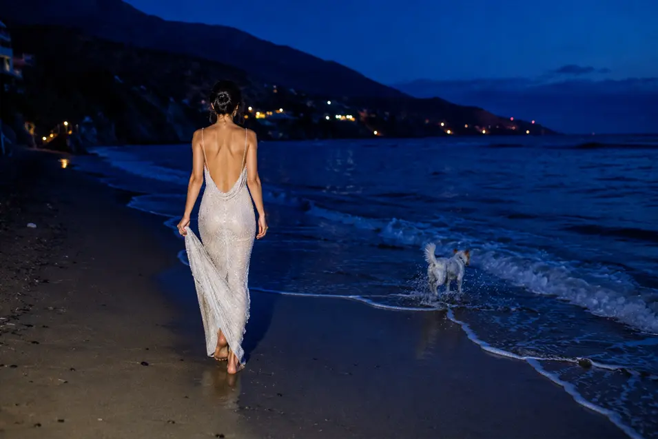 A bride in a slinky pearl-beaded gown walks barefoot along the waterline after sunset, photographed from far behind.