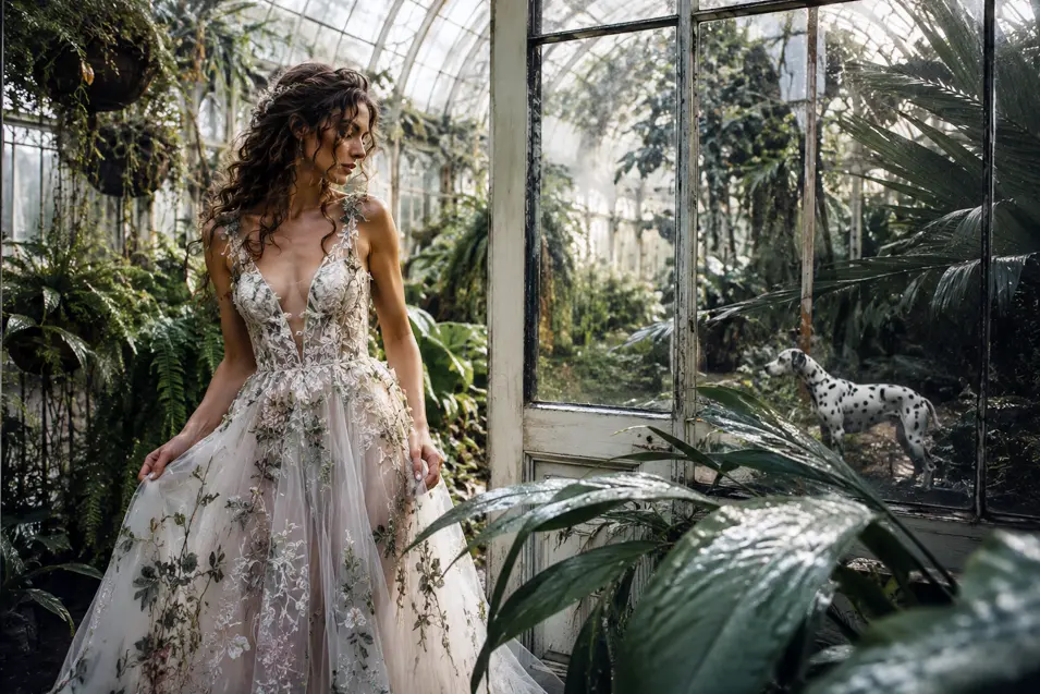 A bride in a botanical lace gown wanders through a lush glasshouse while a Dalmatian explores the tropical greenery nearby.