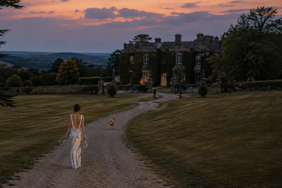 A bride in a sleek white gown follows her dog along a winding drive toward a grand country estate beneath a glowing sunset sky.
