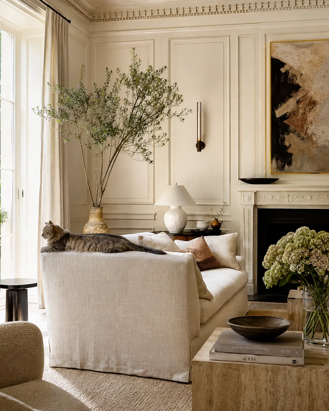 A fluffy grey cat lounges atop a cream linen sofa in an elegant, art-filled living room with classical millwork and warm natural light.