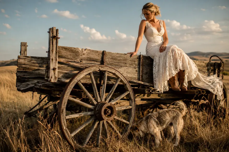 A bride in a lace gown and cowboy boots sits on a weathered wagon while her shaggy dog explores the golden country grass below.