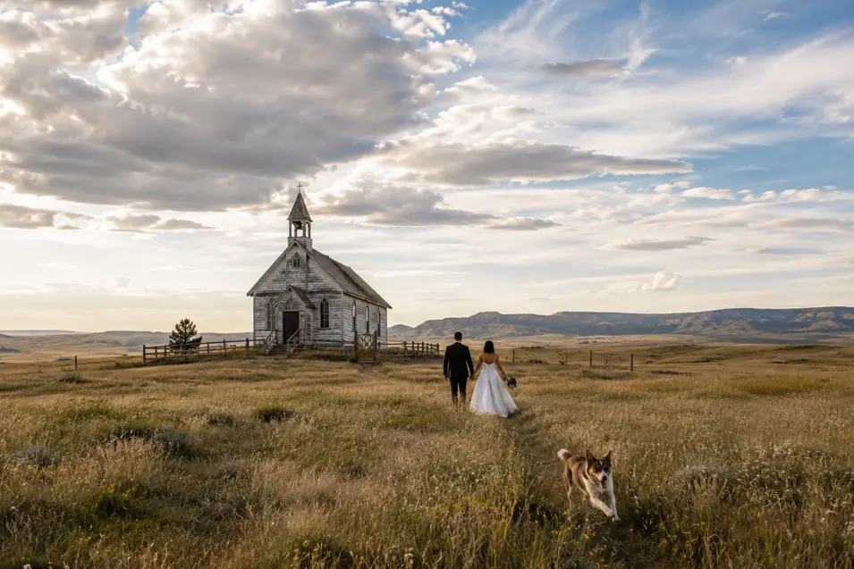 A newlywed couple walks toward a weathered country church across open prairie as their dog runs through the grass nearby.