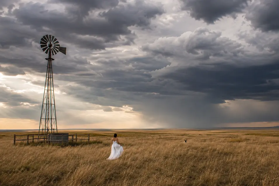 A bride crosses open prairie with her dog beneath dramatic storm clouds, framed by a lone windmill and distant rain.