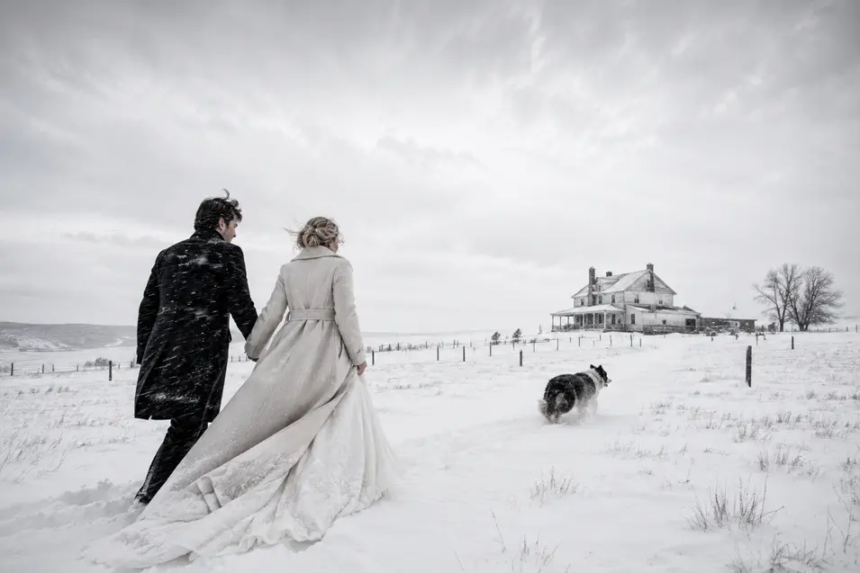 A newlywed couple crosses a windswept snowy field toward an old country farmhouse as their dog bounds ahead through the fresh snow.