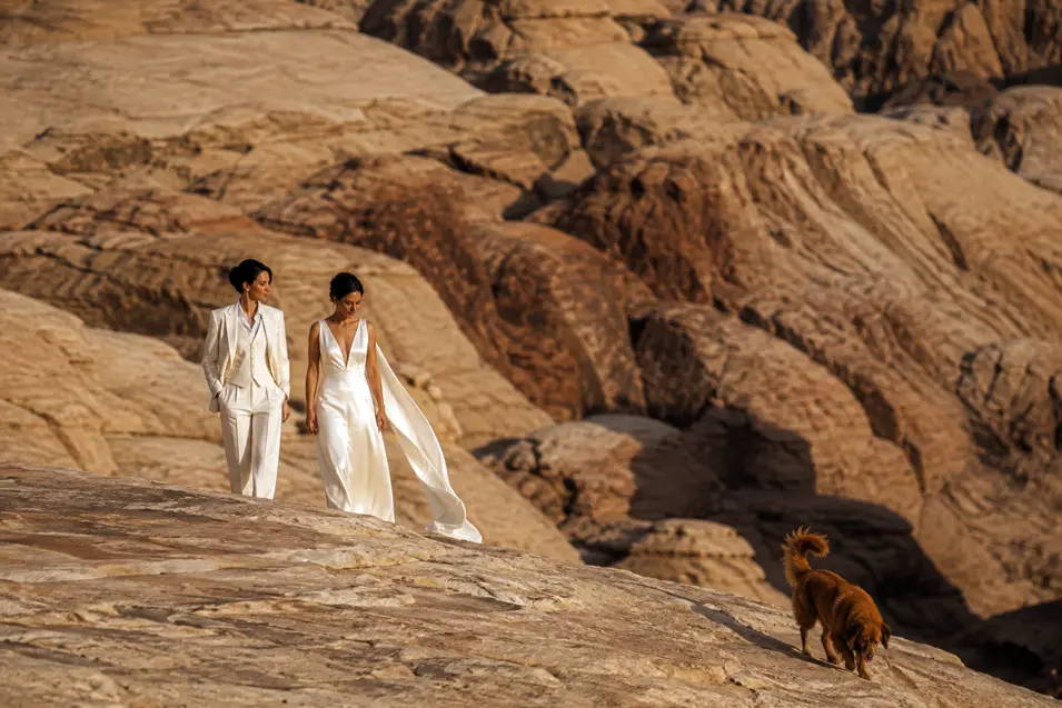 Two brides explore a dramatic desert landscape in elegant wedding attire while their adventurous dog leads the way across the rocks.