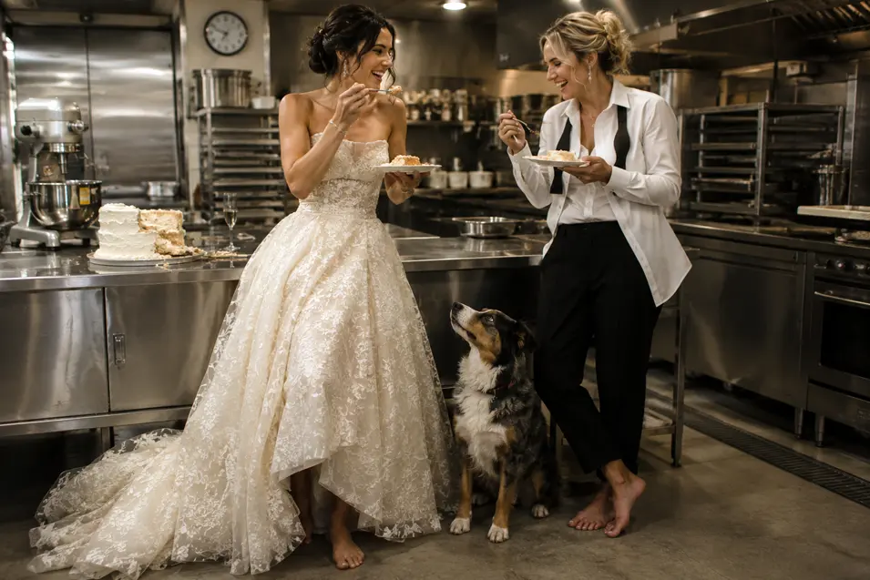 Two brides sneak wedding cake in a restaurant kitchen while their hopeful dog watches closely for a bite.