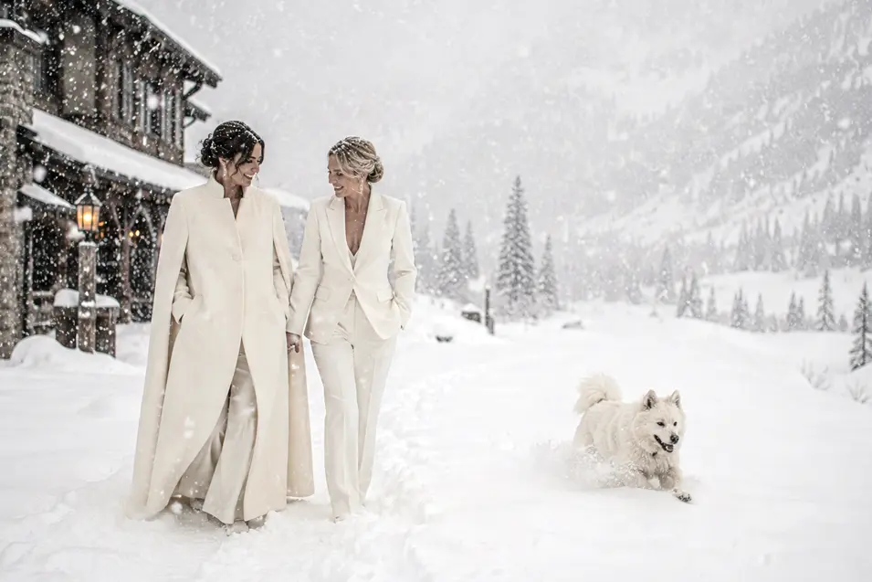 Two brides stroll through deep snow outside a mountain lodge while their playful white dog bounds happily beside them.