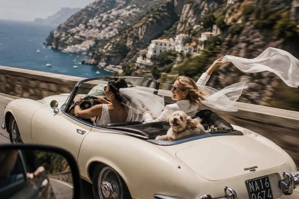 Two brides speed along the Amalfi Coast in a vintage convertible, their little dog happily riding along for the adventure.