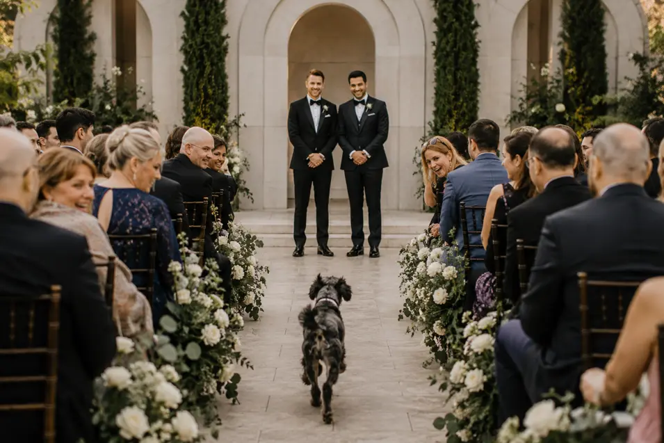 Two grooms smile at the altar as their excited dog runs down the flower-lined aisle toward them.