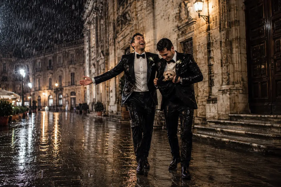 Two grooms laugh in the pouring rain after their wedding, carrying their little dog through a beautifully lit historic city street.