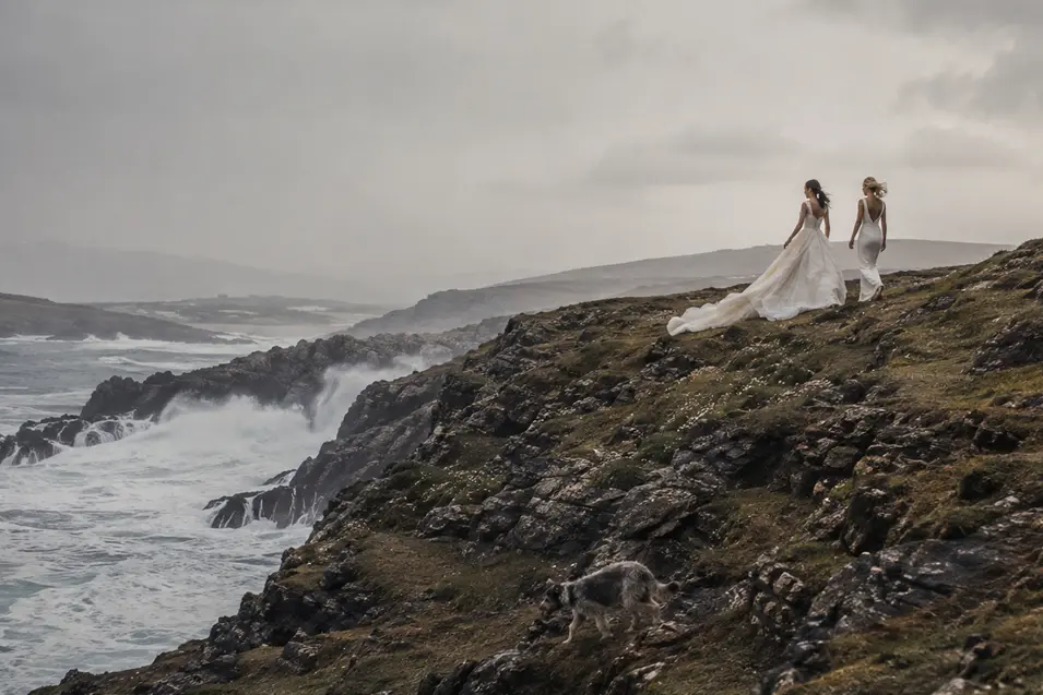 Two brides walk along a wild, windswept coastline as their adventurous dog explores the rocky cliffs below.