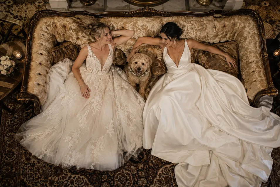 Two brides relax together on an ornate vintage sofa with their happy little dog nestled between them after the wedding.