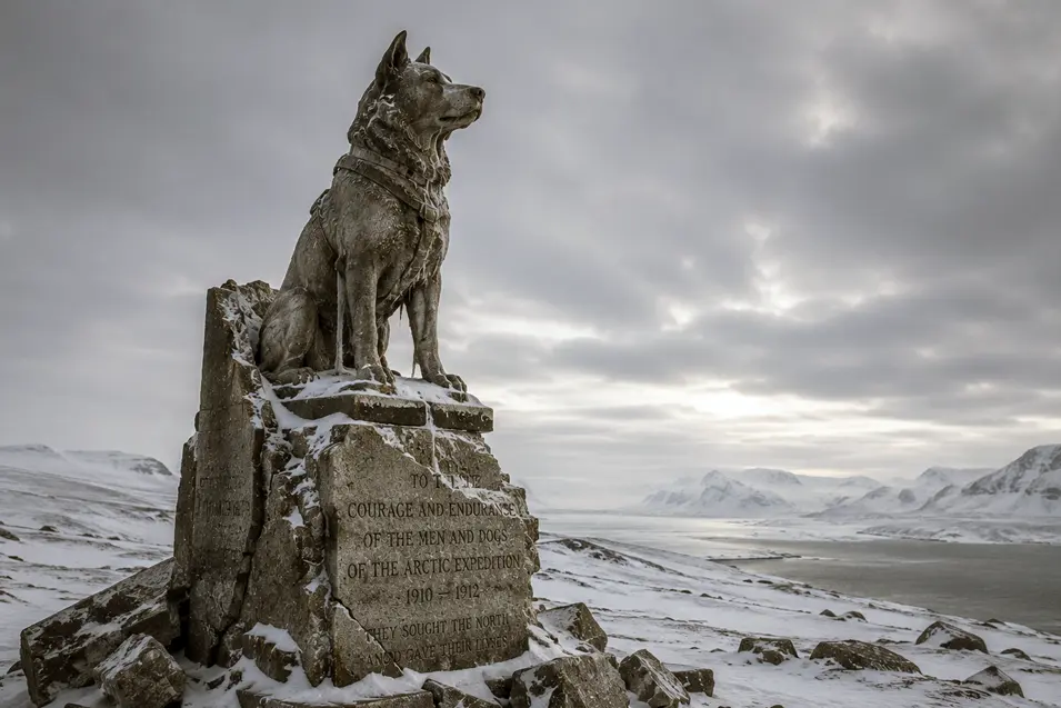 A weathered dog monument honors the courage and endurance of Arctic explorers against a vast, snow-covered polar landscape.
