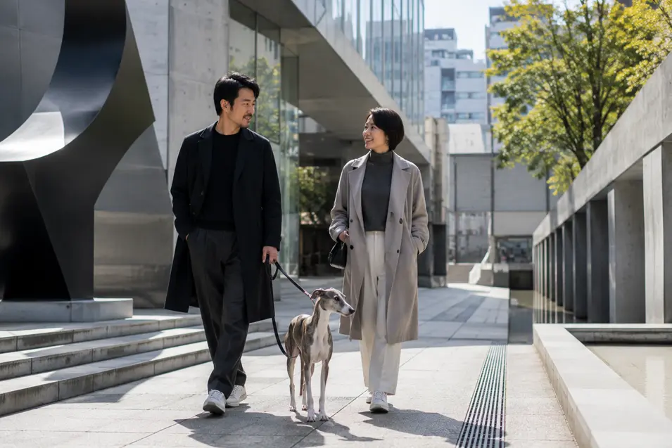 A stylish couple walks a slender greyhound through a modern Japanese plaza surrounded by striking contemporary architecture.