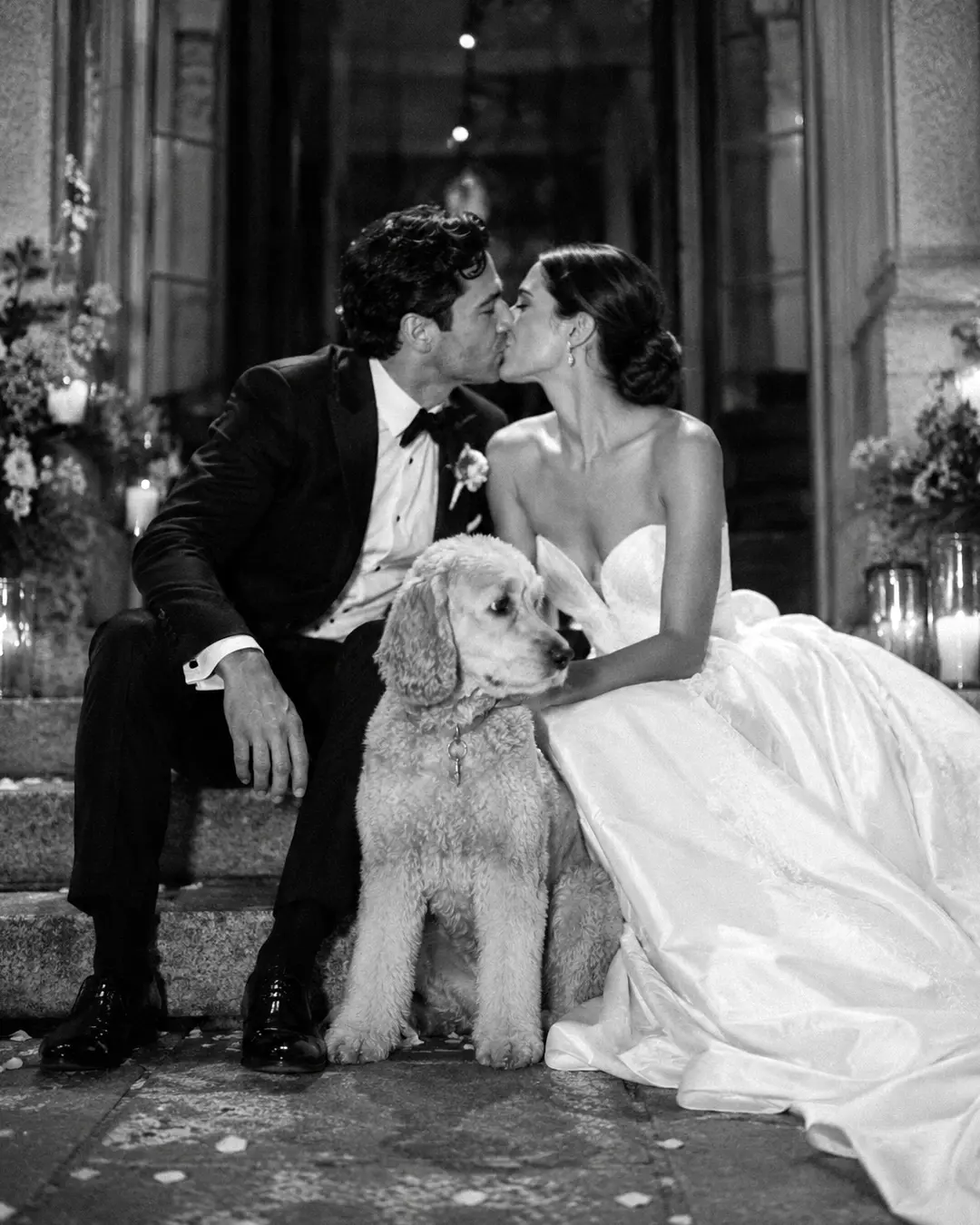 A bride and groom share a kiss on stone steps with their curly-coated dog between them in an elegant black-and-white wedding portrait.