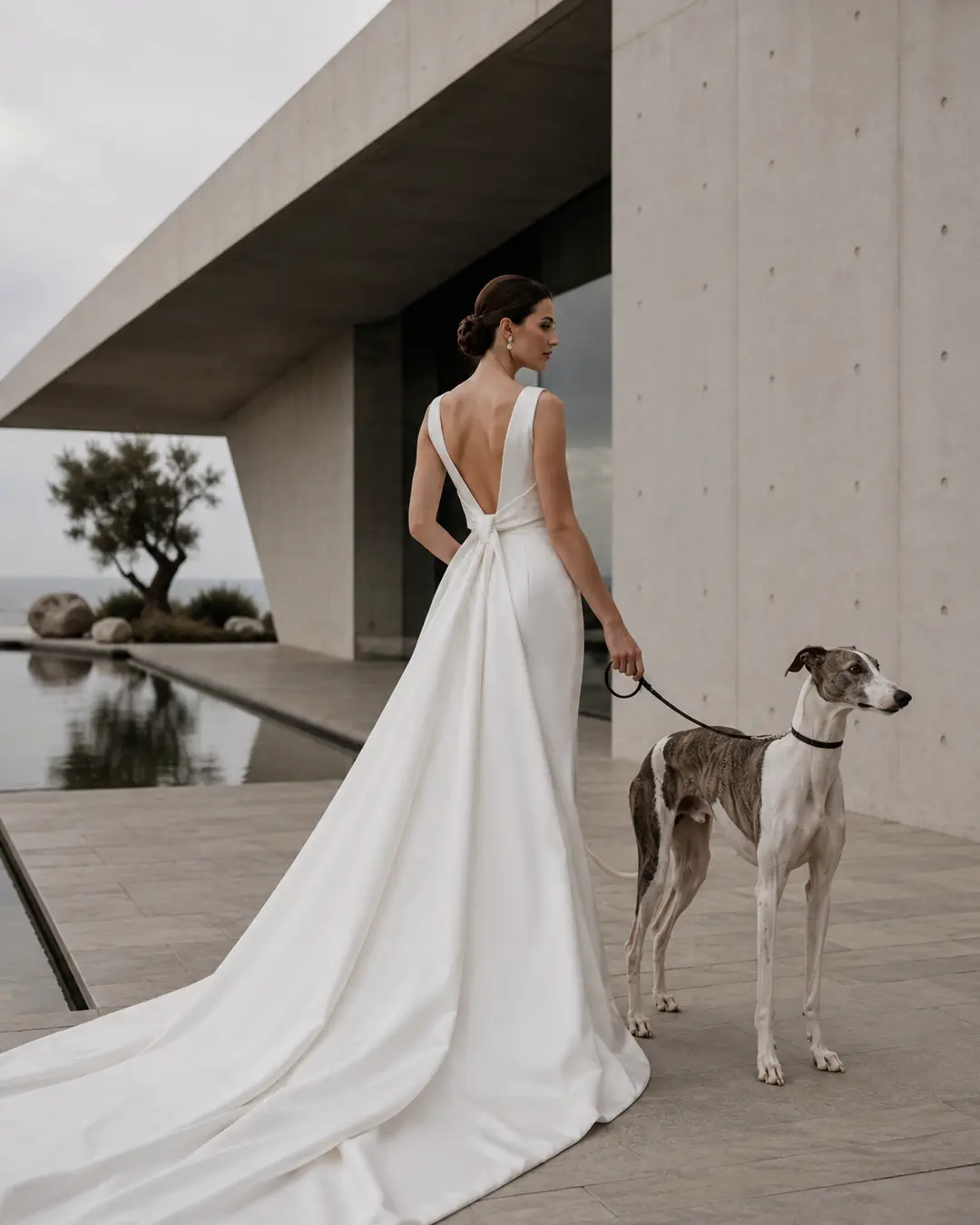 A bride in a sleek white gown walks a brindle Greyhound beside a reflecting pool at a striking minimalist concrete wedding venue.