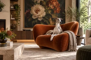 A Whippet sits elegantly in a sculptural rust velvet chair, surrounded by dramatic floral art, warm wood and lush greenery.